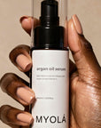 Hand holding a bottle of MYOLA argan oil serum against a beige background
